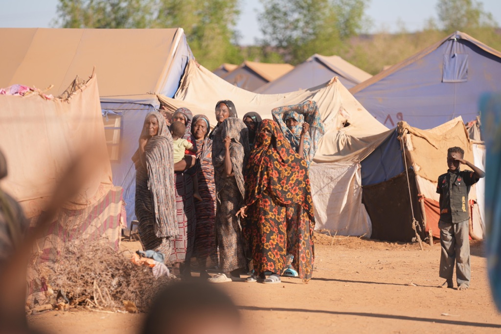 Zelte (Flüchtlingscamp?), eine Gruppe Frauen im lokalen Stil gekleidet, z.T. verschleiert, eine mit Kleinkind im Arm, mit etwas Abstand ein Jugendlicher, alle mit Blick zur Kamera